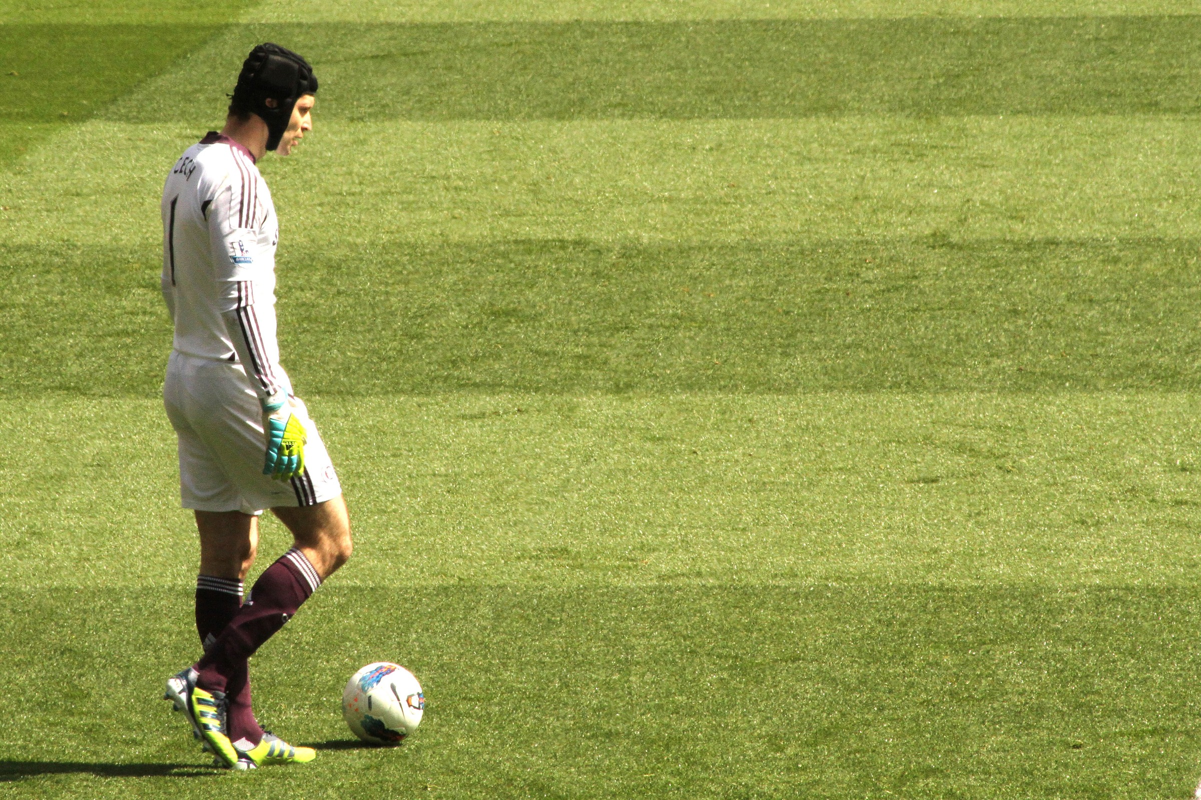 Petr Čech in a grey Chelsea goalkeeper kit and protective headguard standing over the ball on the pitch in 2012.